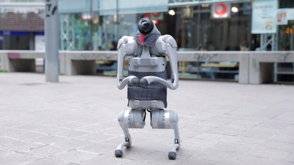 Un robot bípedo de color gris claro de pie en una plaza peatonal. El robot tiene una estructura mecánica articulada y lleva puesto un chaleco o prenda de tela vaquera oscura alrededor de su torso.