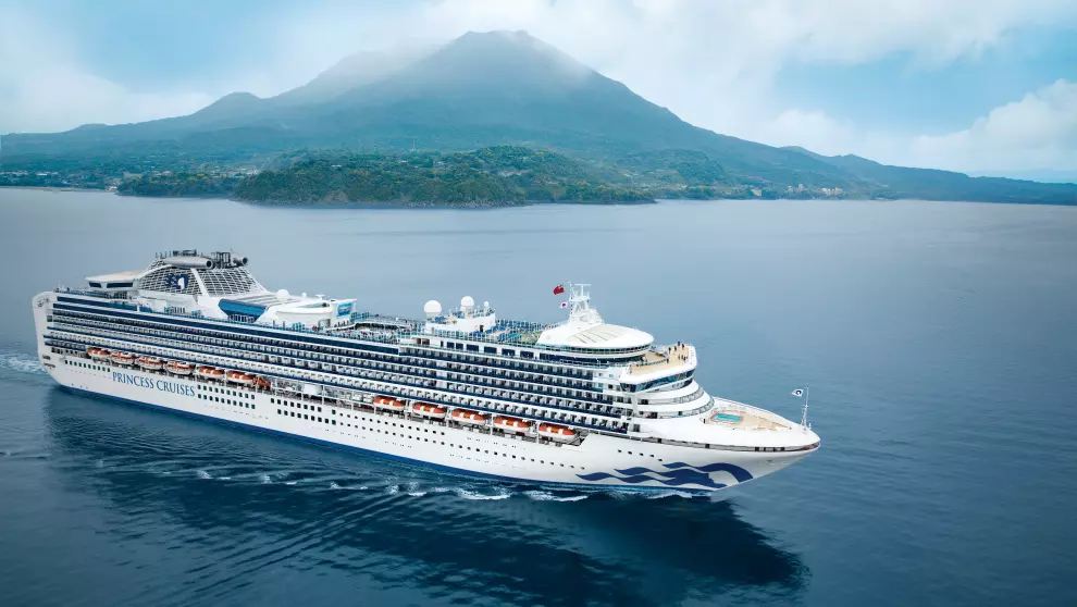 Vista panorámica del crucero Diamond Princess navegando por aguas tranquilas con una majestuosa montaña envuelta en nubes al fondo.