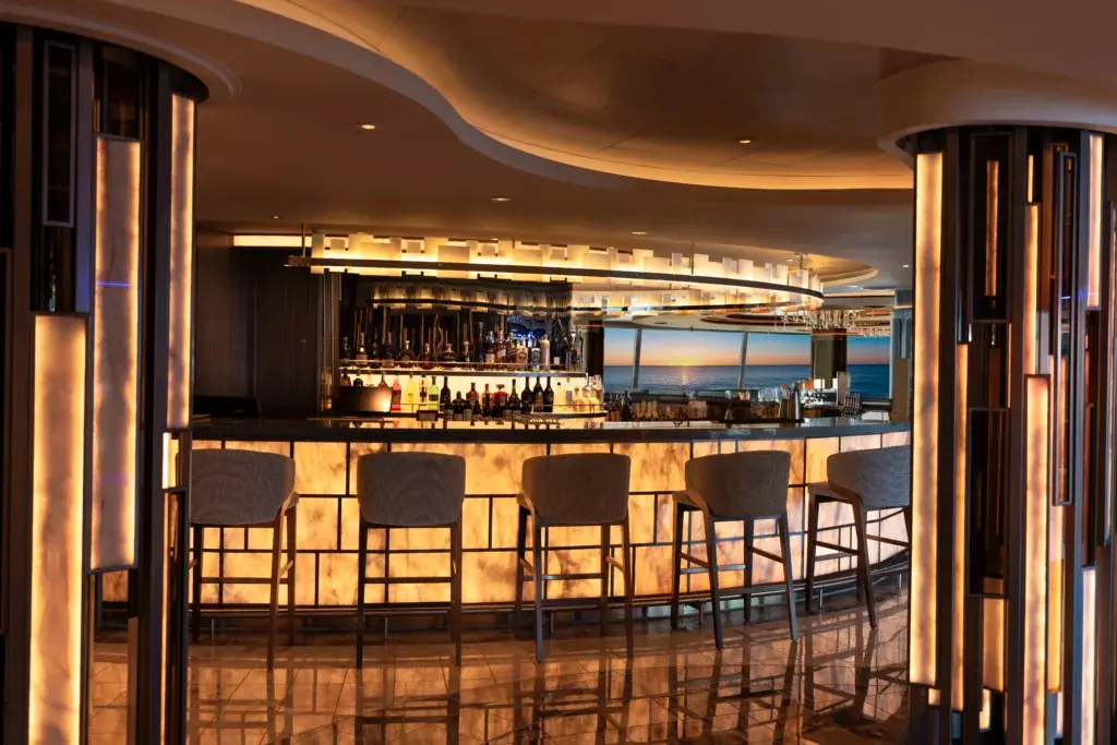 Elegante bar de diseño a bordo del buque Allura de Oceania Cruises con iluminación cálida y vistas panorámicas al océano durante el atardecer.