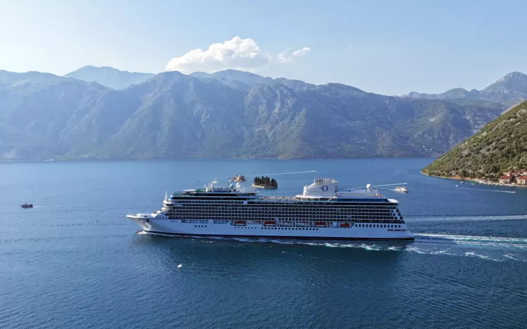 Vista lateral del nuevo barco Allura de Oceania Cruises navegando por aguas cristalinas frente a una costa montañosa en un itinerario europeo.