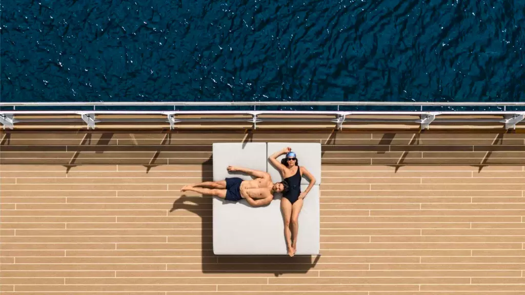Pareja descansando de forma relajada sobre una cama de día en la cubierta exterior de madera frente al mar azul