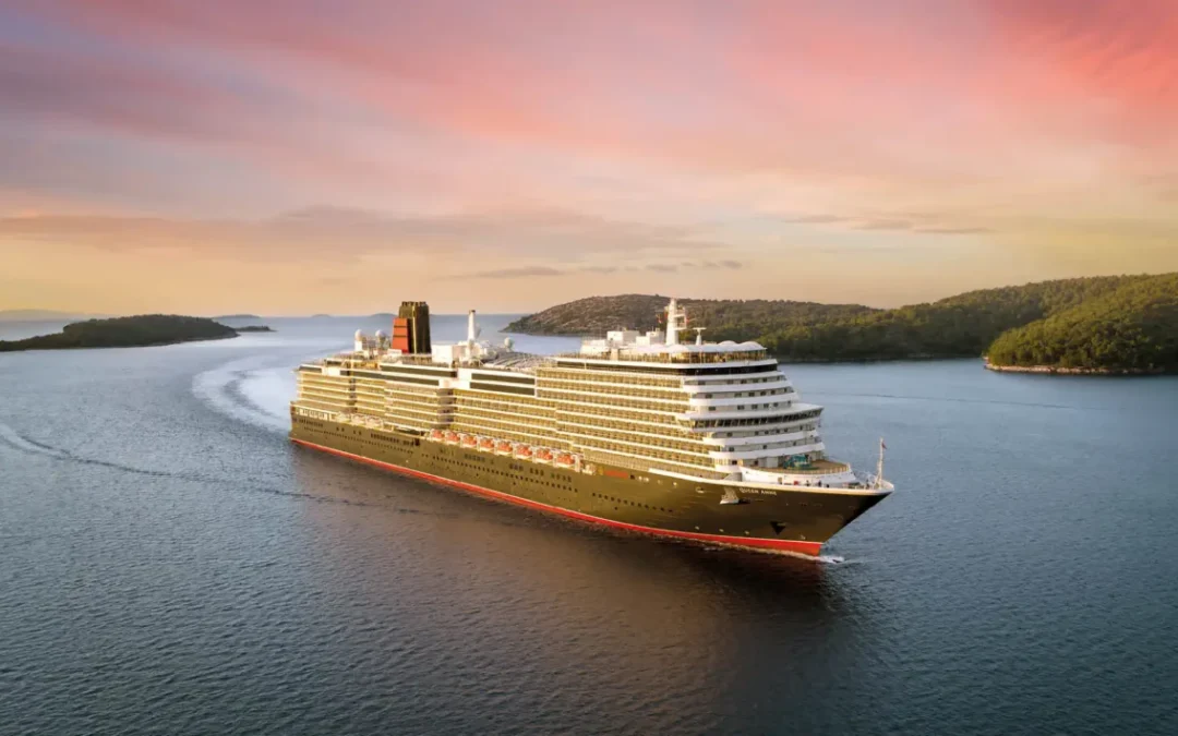 Vista aérea del barco de cruceros Cunard Queen Anne navegando por una bahía costera al atardecer, con su distintiva chimenea roja y negra.