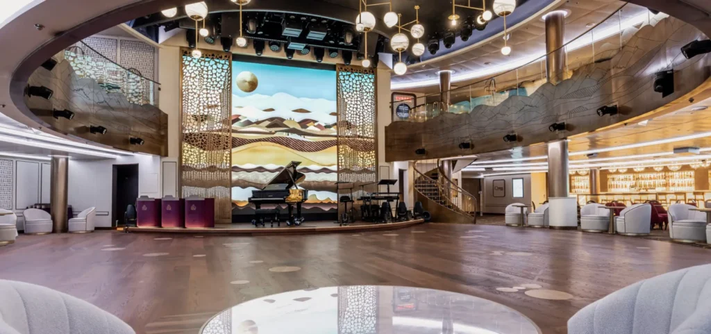 Pista de baile de madera en el salón Queens Room del barco Cunard Queen Anne, con un piano de cola en el escenario y un mural de paisaje iluminado al fondo.