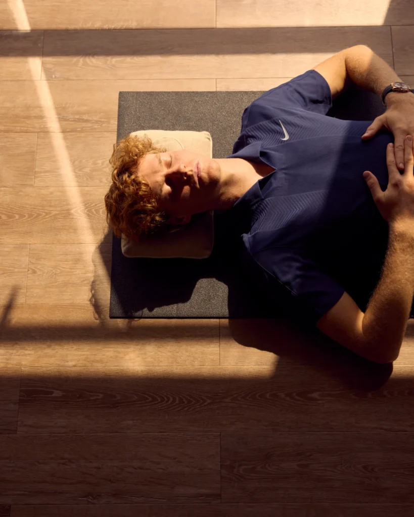Jannik Sinner acostado en una esterilla de yoga en un gimnasio de crucero, mirando hacia el horizonte infinito sobre el océano