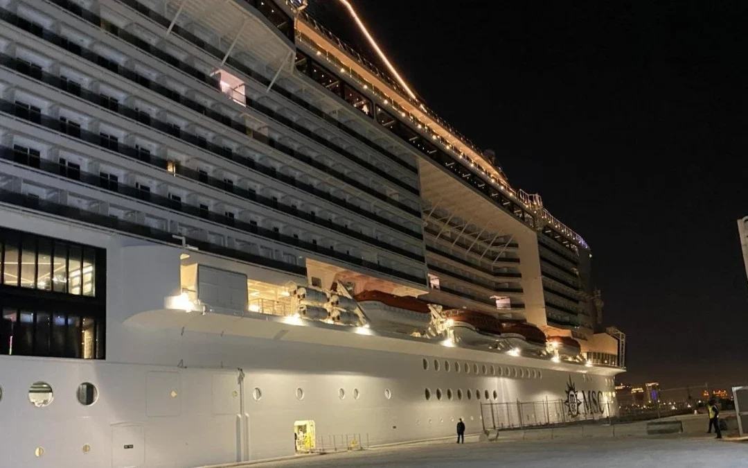 Vista nocturna detallada de un crucero de MSC Cruises atracado en un muelle portuario. El barco está fuertemente iluminado, mostrando múltiples cubiertas de balcones, botes salvavidas y una gran pared de vidrio en la parte inferior. Personal del puerto y un edificio de terminal moderno a la derecha.