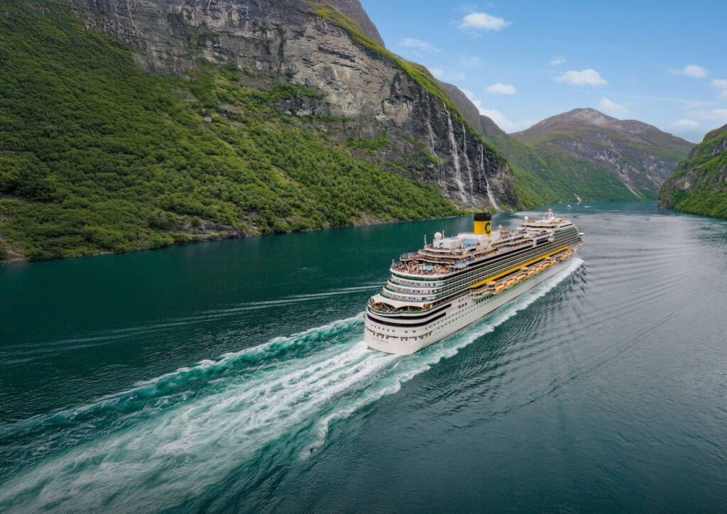 El barco Costa Diadema navegando por las aguas verdes del fiordo de Geiranger en Noruega, rodeado de altas montañas y cascadas.