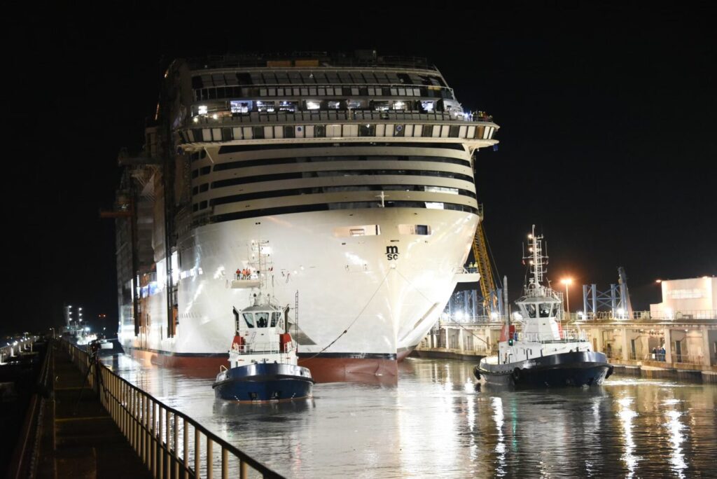 El crucero MSC World Europa siendo remolcado por dos barcos en un astillero durante la noche.