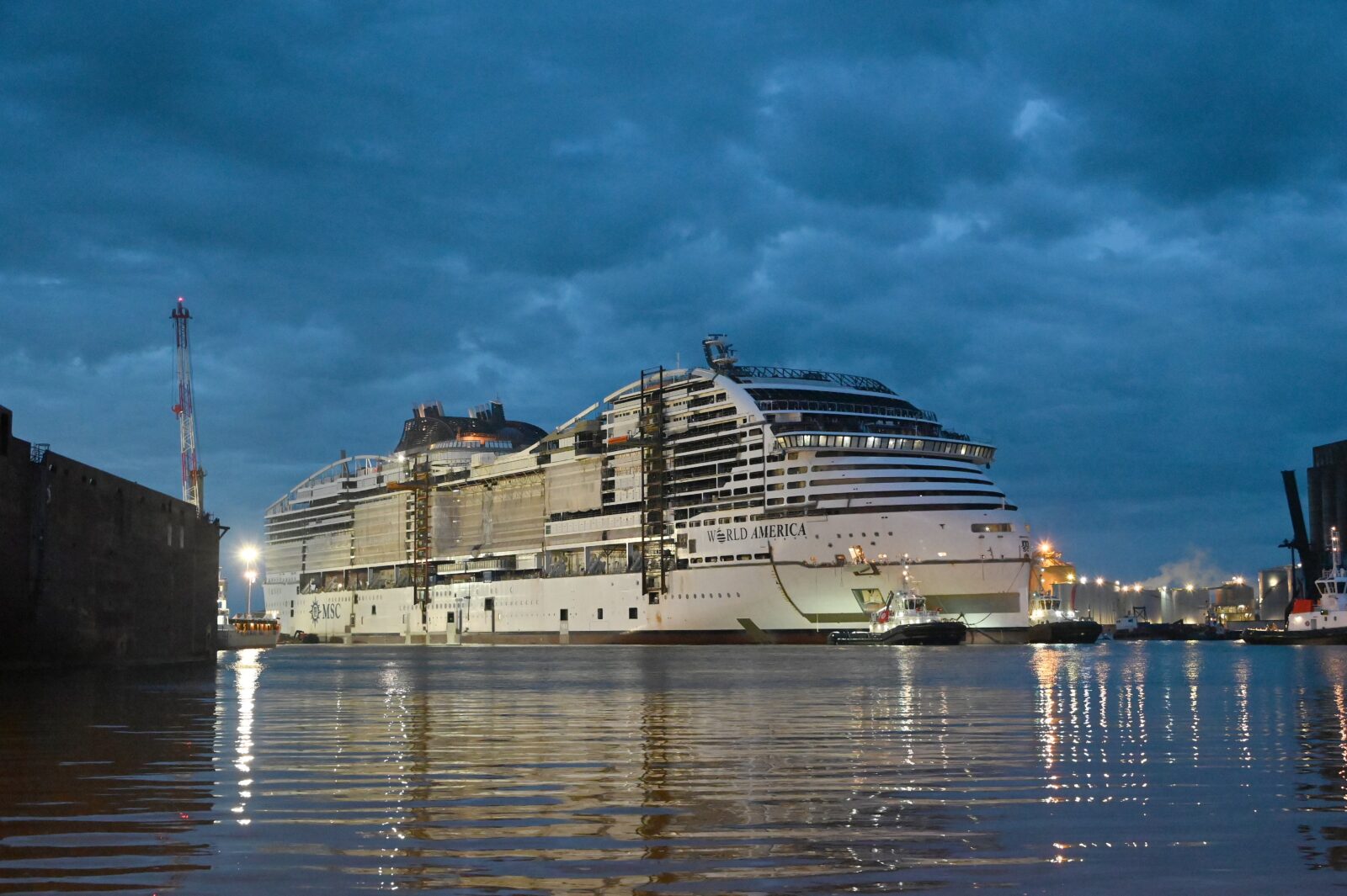 El crucero MSC World America en construcción en un astillero durante el atardecer, rodeado de remolcadores.