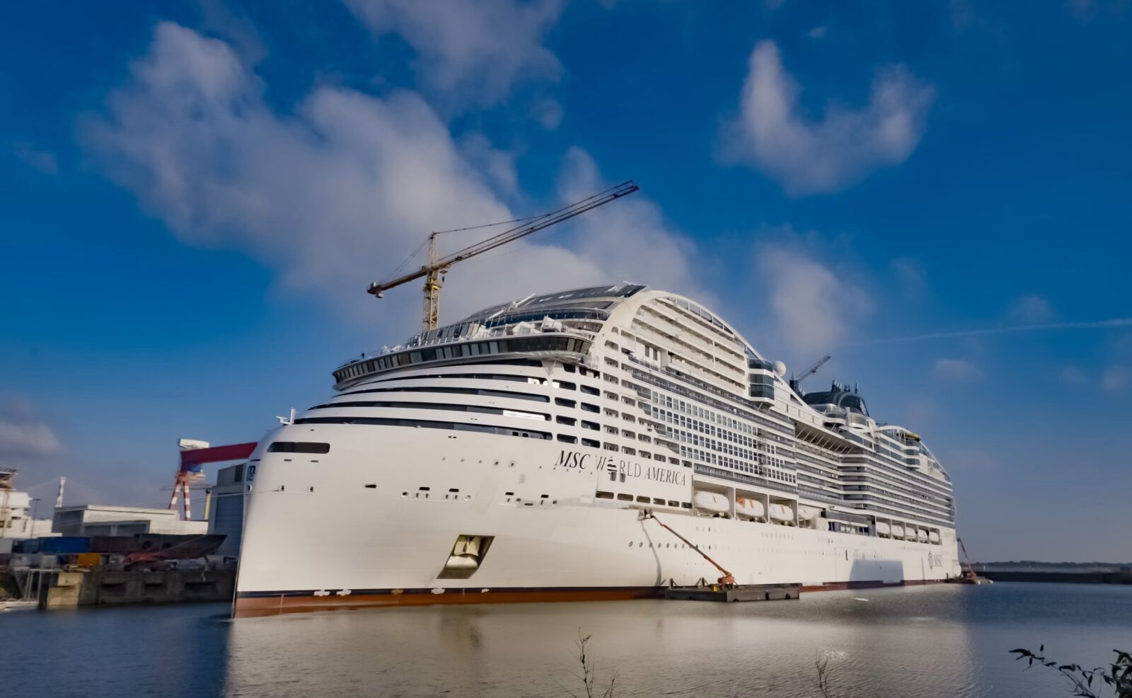 El crucero MSC World America en fase de construcción atracado en el astillero bajo un cielo azul con una grúa visible.