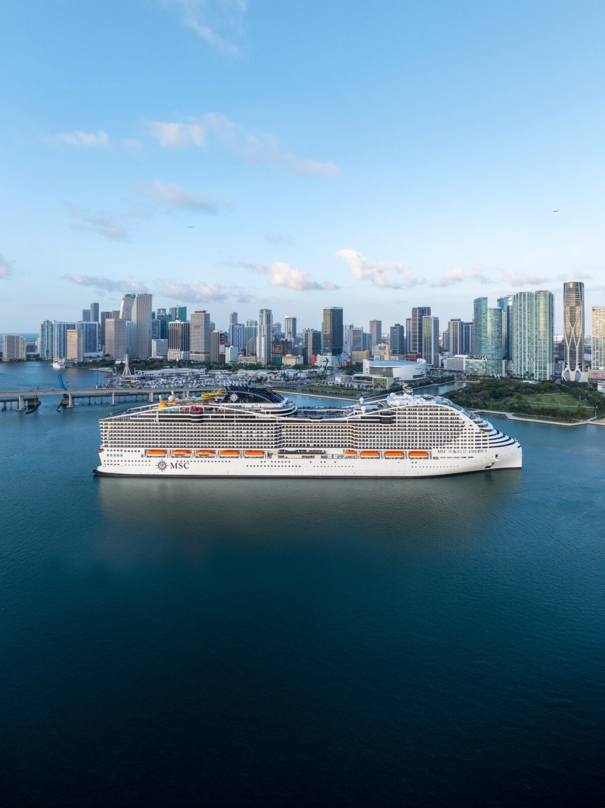 El crucero MSC World America navegando frente a la bahía y el horizonte de rascacielos de Miami al atardecer.