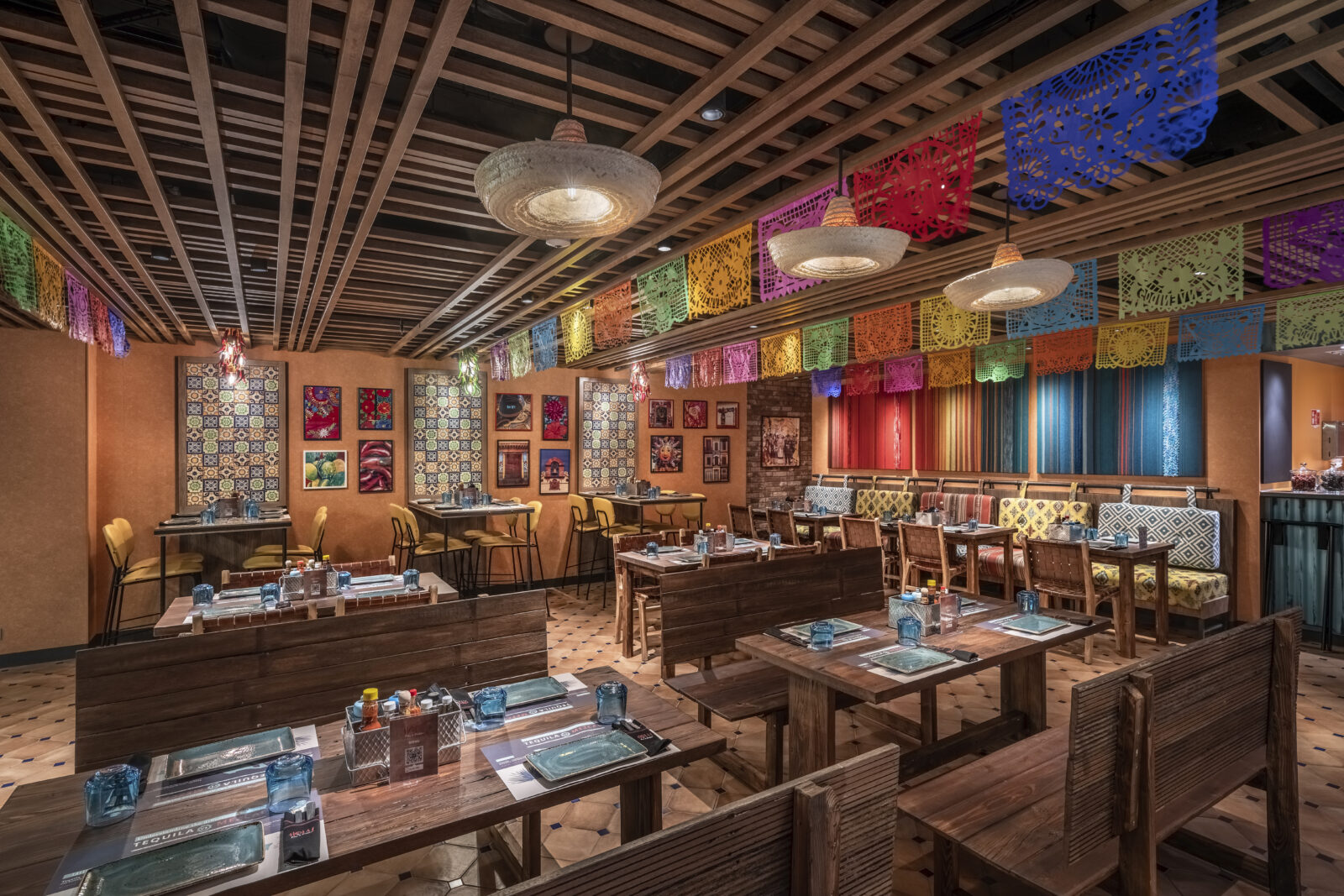 Interior de restaurante con mesas de madera, bancos y decoración mexicana con papel picado colorido en el techo.