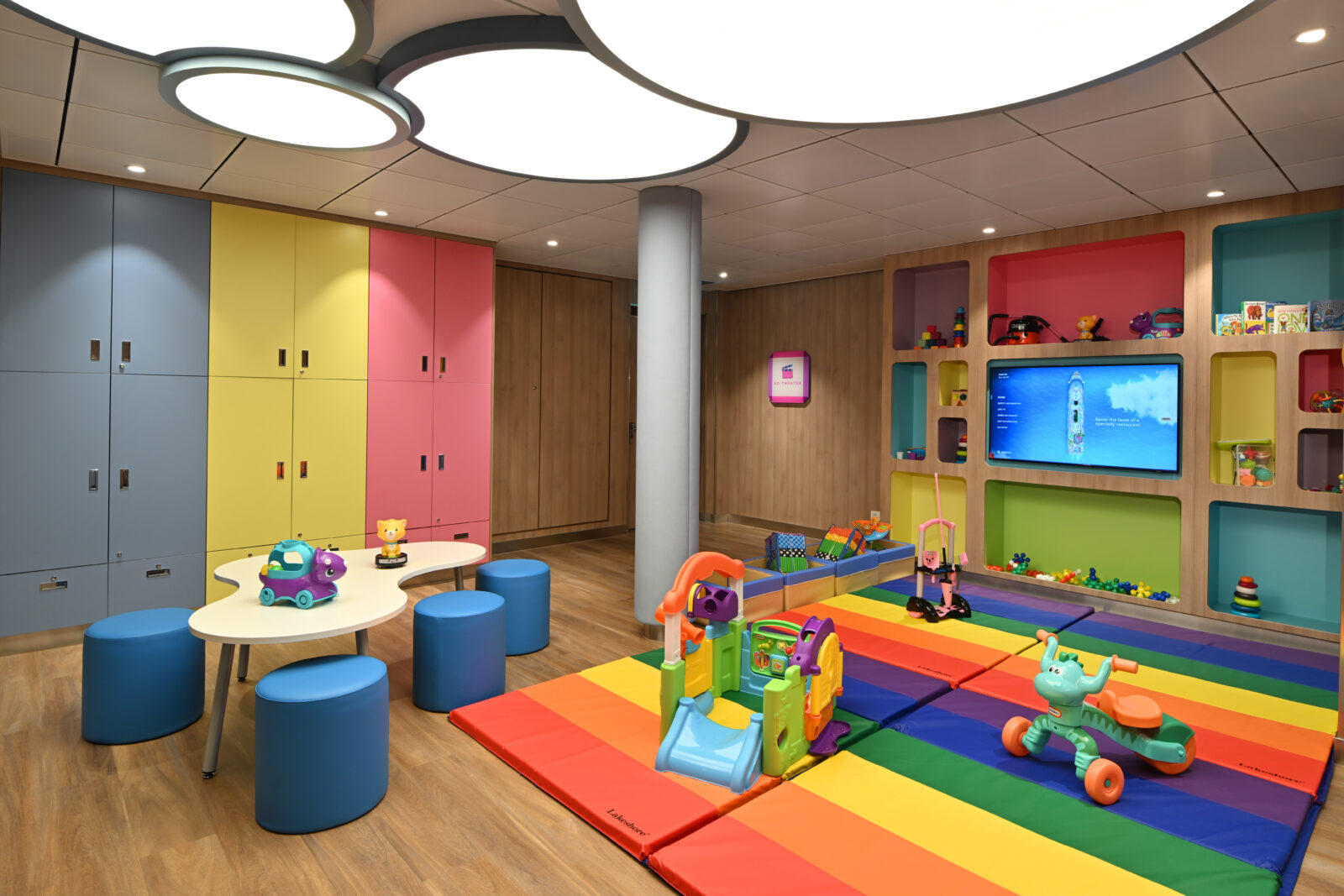 Sala de juegos infantil con casilleros de colores, mesa con taburetes, alfombra de arcoíris y juguetes diversos