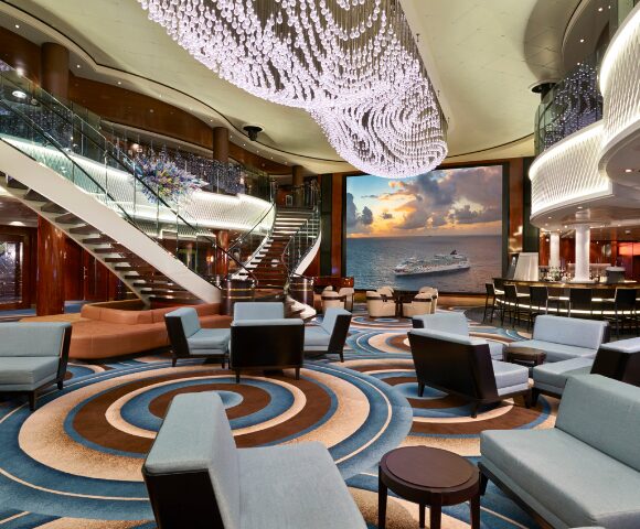 Vista del atrio central del crucero Norwegian Gem con una gran lámpara de araña, escaleras curvas y una pantalla gigante que muestra un barco en el mar.
