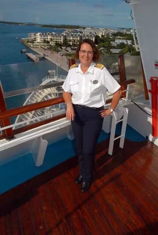 Una oficial con uniforme de capitana de crucero posa en la cubierta exterior de un barco con vistas a un puerto costero.
