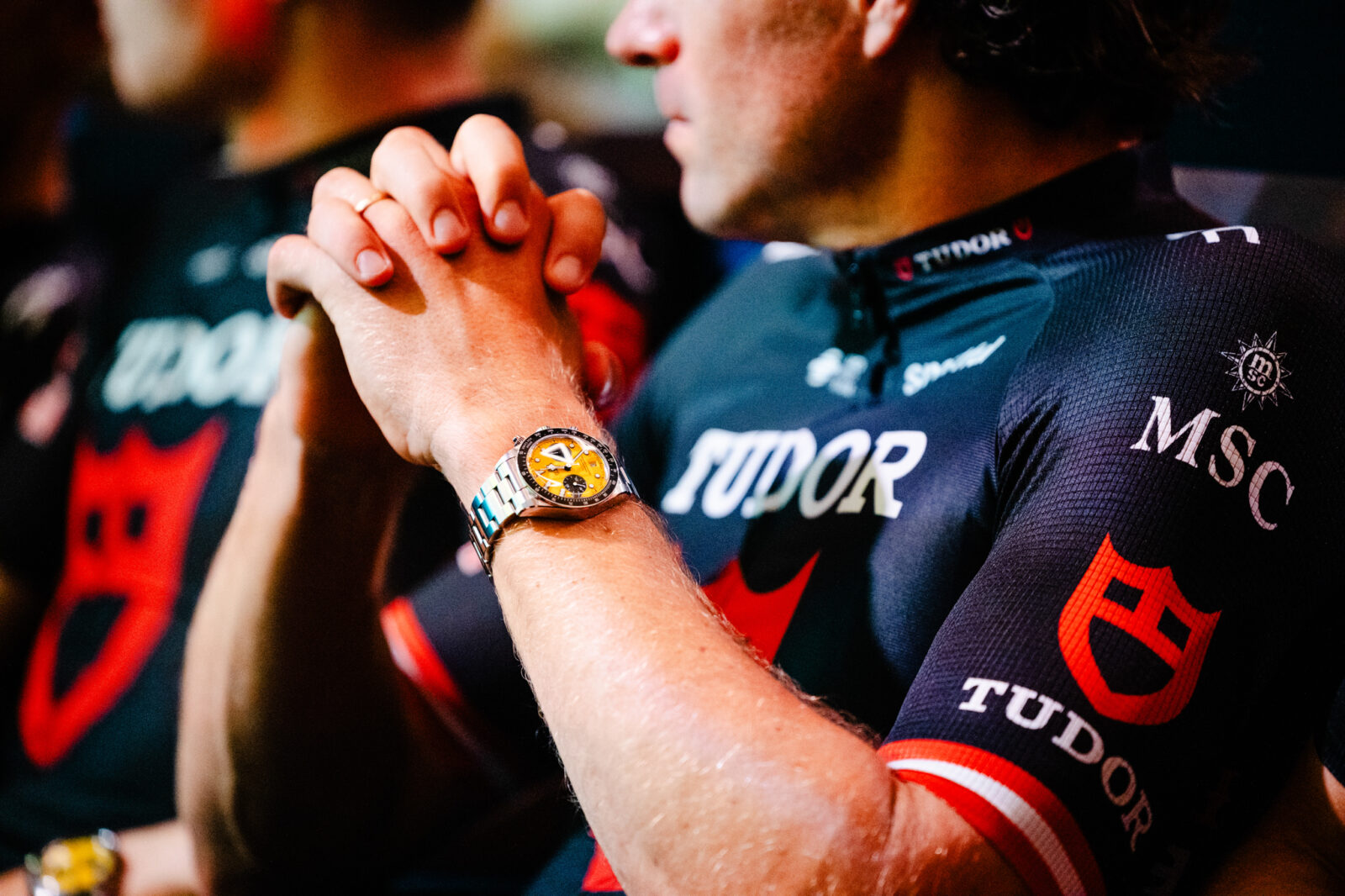 Ciclista con las manos entrelazadas luciendo un reloj de pulsera Tudor con esfera amarilla, vistiendo maillot de equipo.