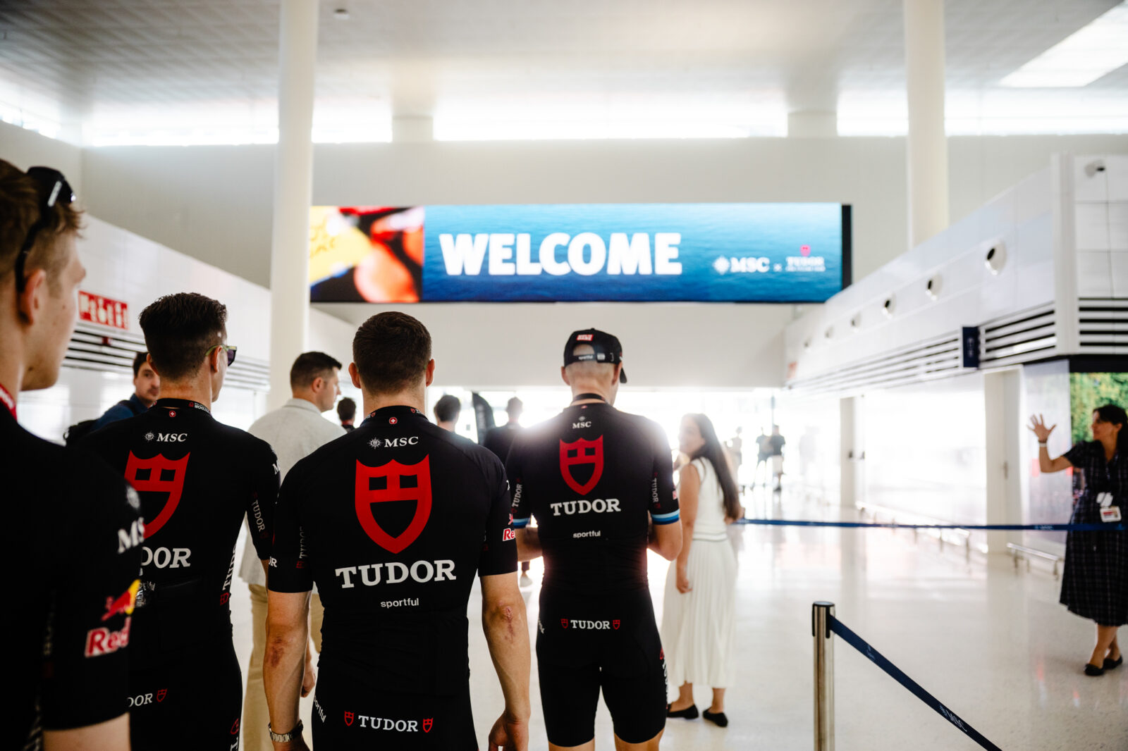 Ciclistas del equipo Tudor de espaldas caminando hacia una pantalla grande que dice WELCOME MSC en una terminal.