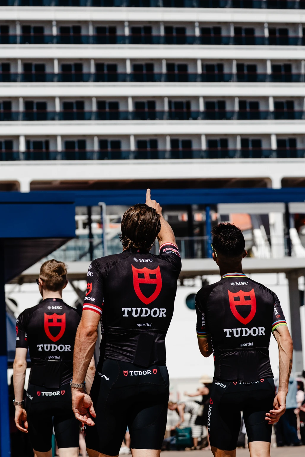 Tres ciclistas del equipo Tudor con uniformes negros y rojos observan un enorme crucero MSC en el puerto.