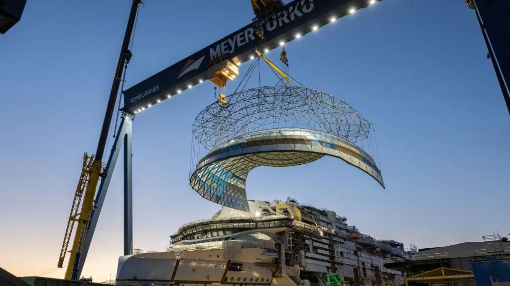 Una grúa pórtico de Meyer Turku eleva una gran estructura metálica curva sobre el casco en construcción del crucero Hero of the Seas.