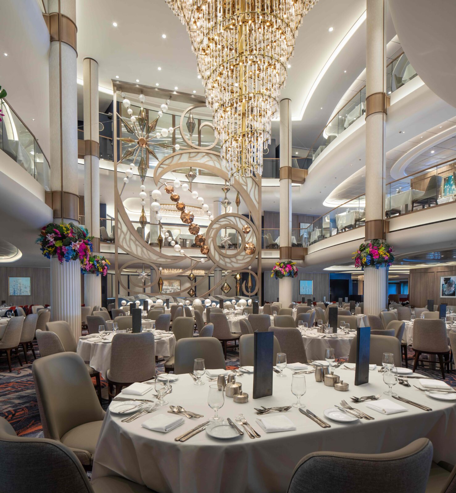 Elegante comedor de varias plantas en un crucero con mesas redondas, sillas beige y una gran lámpara de araña central.