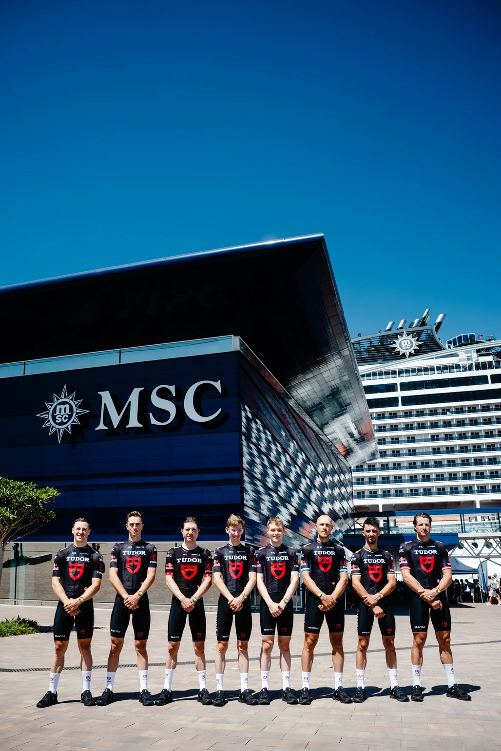Ocho ciclistas del equipo Tudor con uniformes negros posan frente a la terminal de cruceros MSC en Barcelona.
