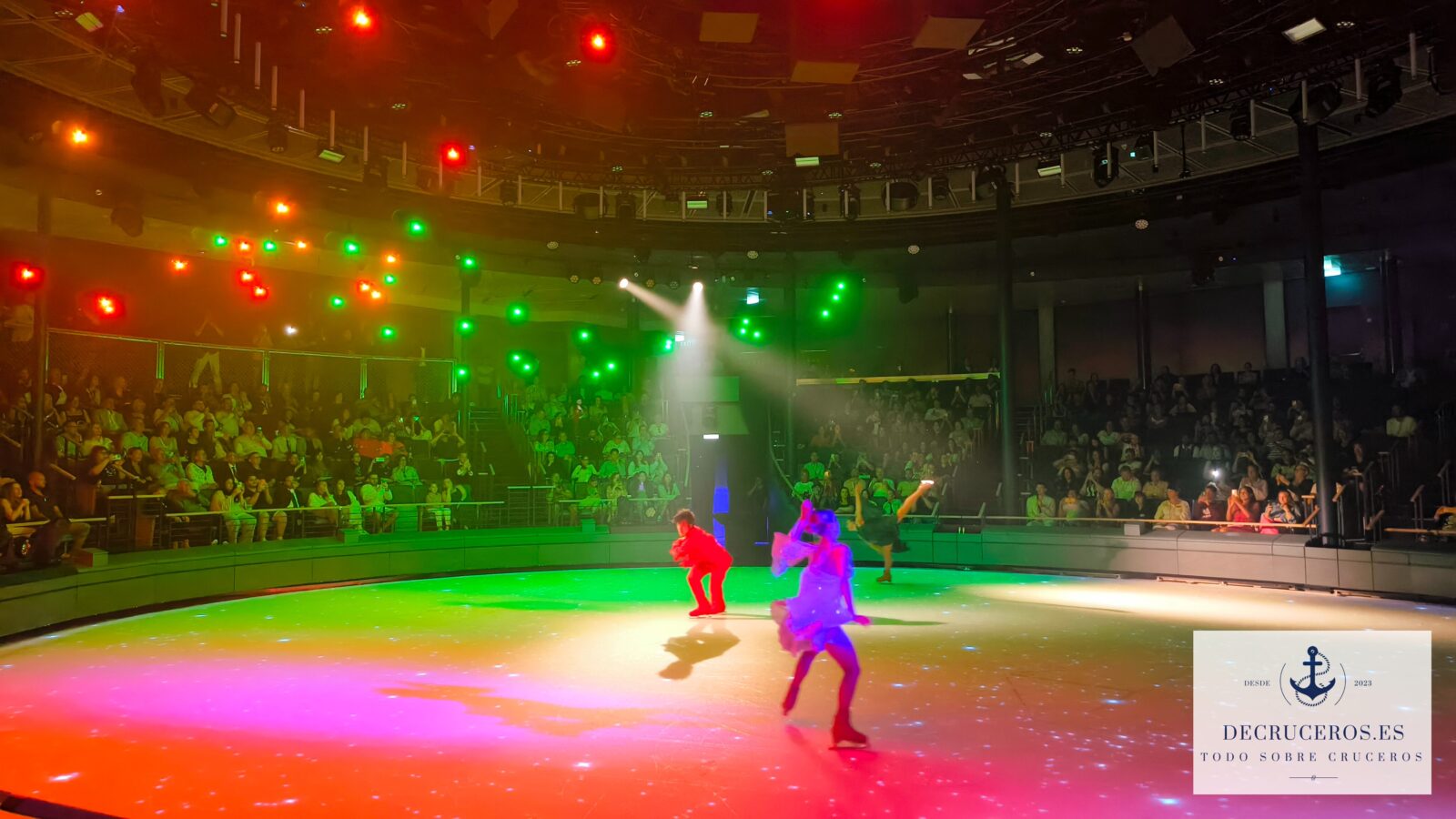 Patinadores sobre hielo realizando una coreografía en una pista circular con iluminación de colores y público de fondo.