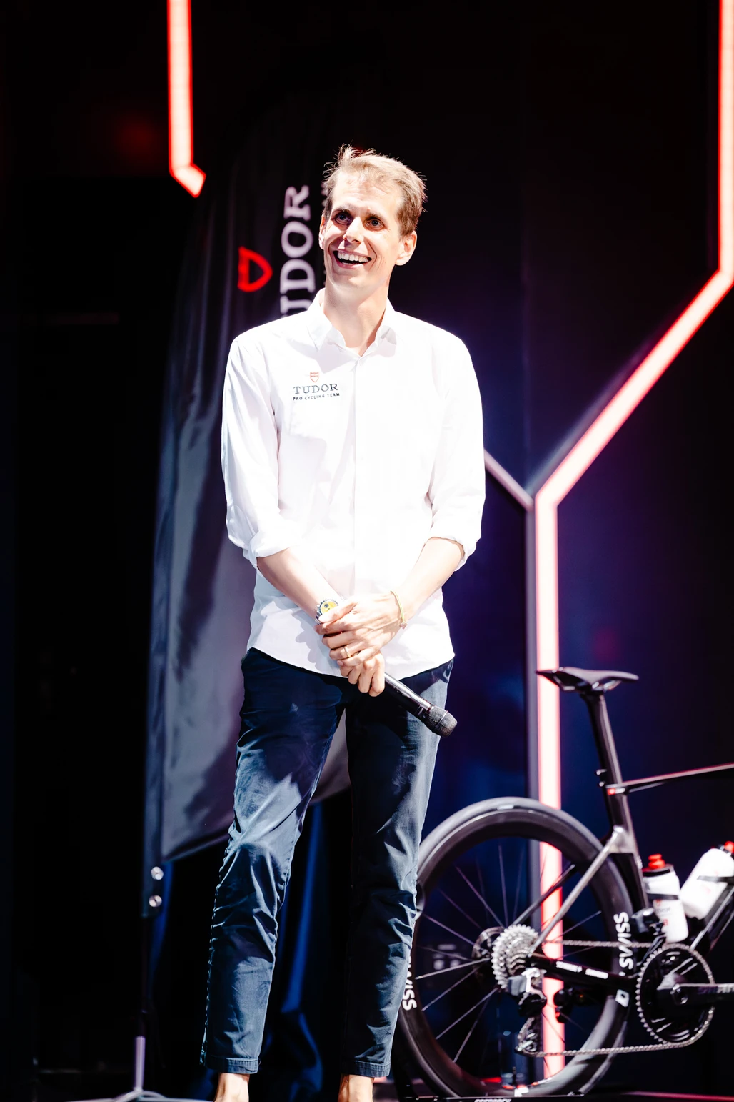 Fabian Cancellara sonriendo en el escenario junto a una bicicleta de competición en un evento del Tudor Pro Cycling Team.