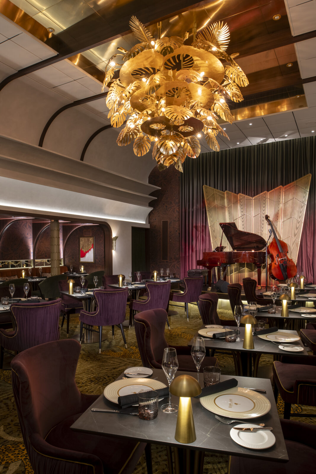 Restaurante elegante con sillas de terciopelo morado, una gran lámpara dorada y un escenario con piano y contrabajo