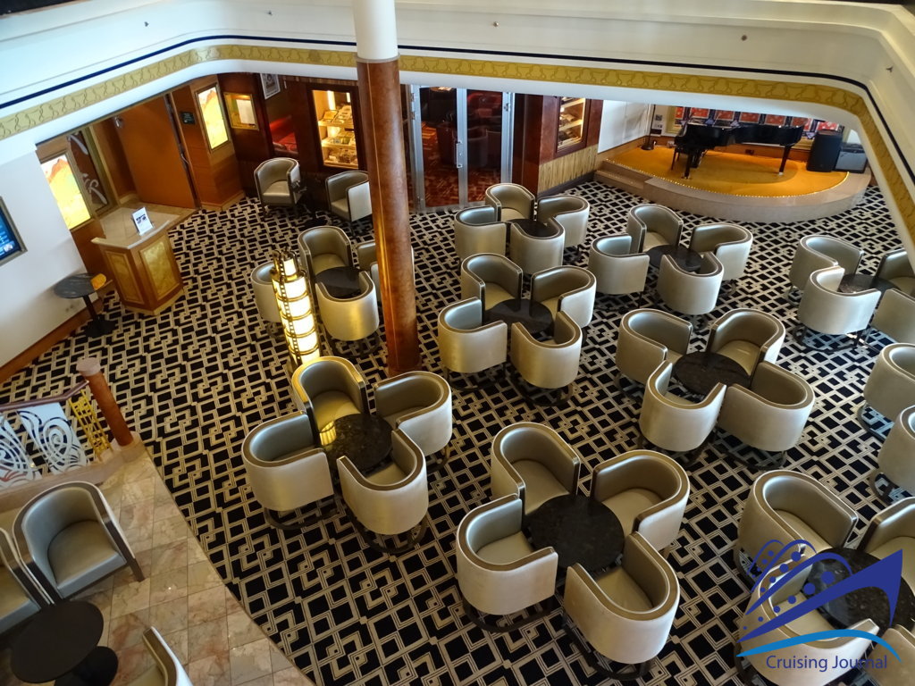 Salón de crucero con moqueta geométrica, mesas redondas con sillones en forma de trébol y un piano de cola al fondo