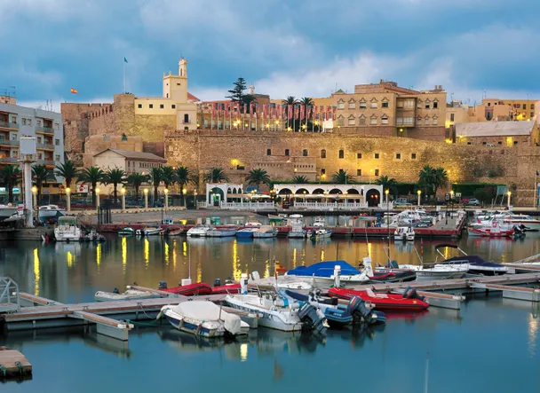 Puerto deportivo de Melilla con embarcaciones en el muelle y murallas de la ciudad antigua al fondo bajo un cielo azul.