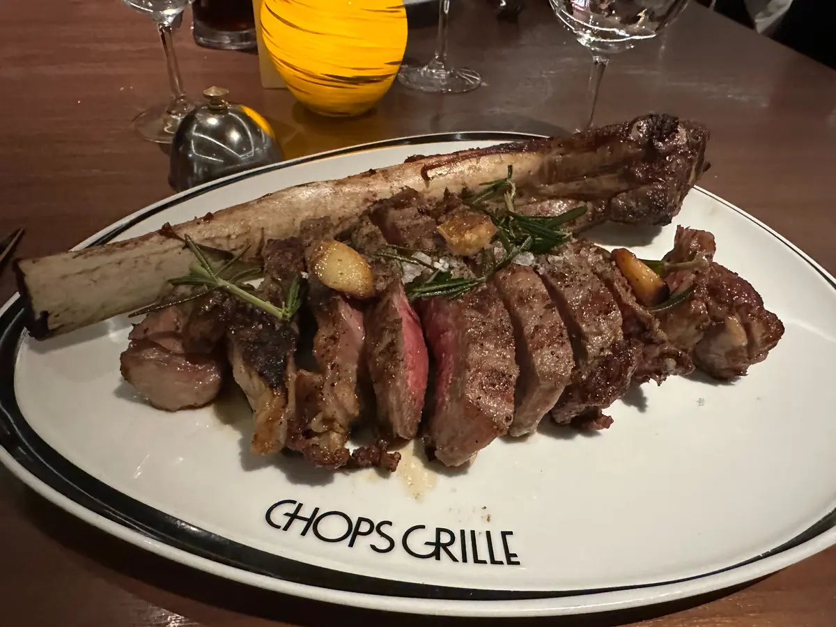 Un corte de carne tomahawk rebanado servido en un plato blanco con el logo de Chops Grille en un restaurante.
