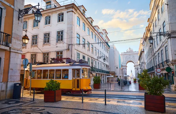 Tranvía clásico amarillo de Lisboa circulando por una calle adoquinada con edificios históricos y un arco triunfal.