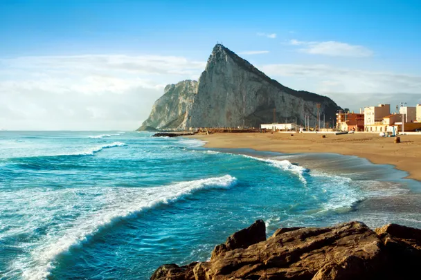 Vista panorámica del Peñón de Gibraltar desde la costa con mar azul y playa de arena.