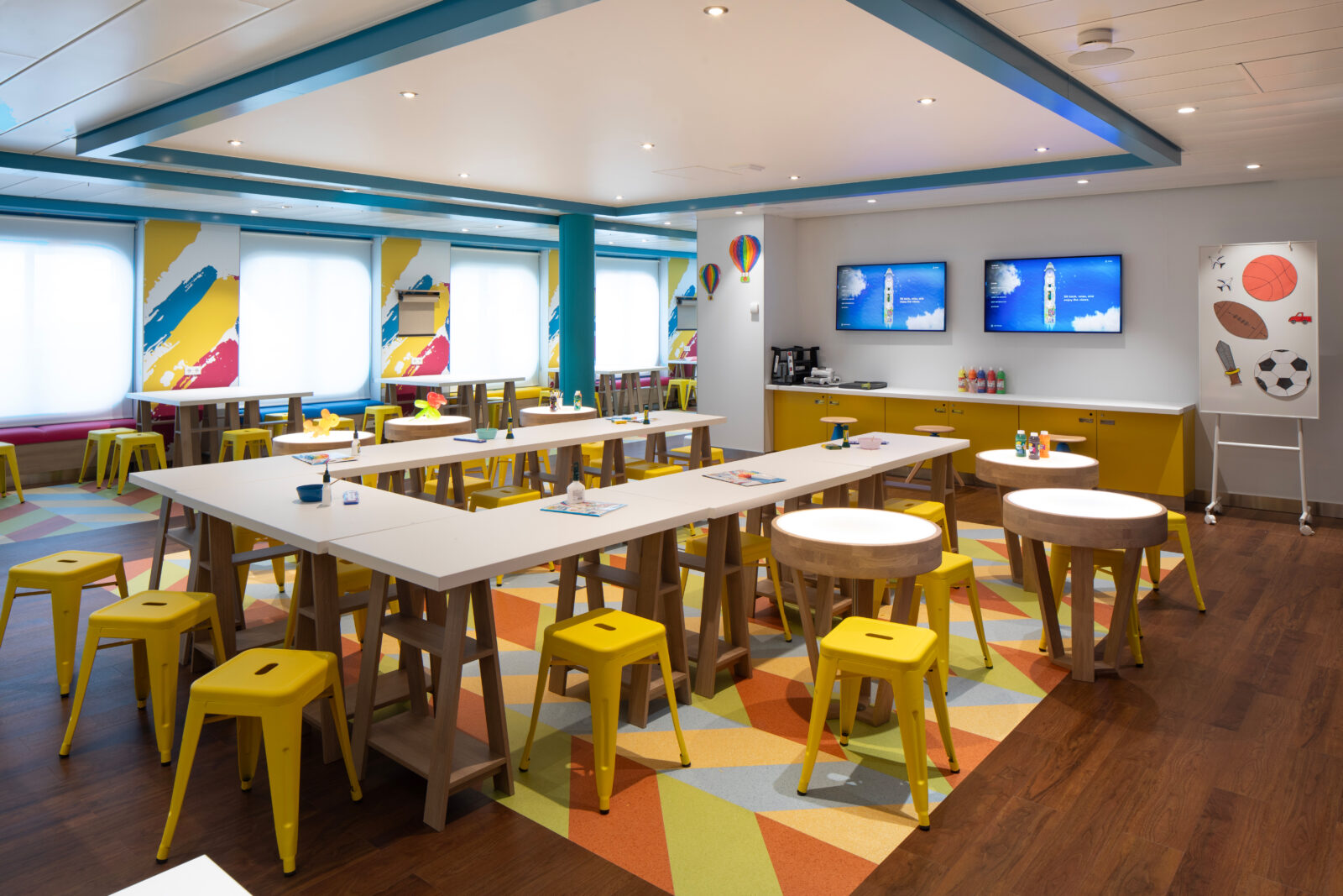 Zona de juegos infantil con mesas blancas, taburetes amarillos y decoración colorida en un crucero.